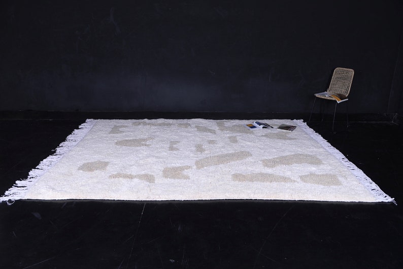May include: White shag rug with a geometric pattern in a lighter shade of beige. The rug has a fringe border and is photographed on a black surface.