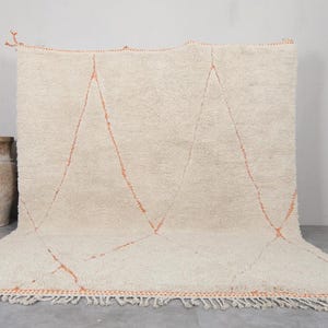 May include: Cream-colored, hand-woven rug with a geometric design. The rug features three large, orange-red diamond shapes. The edges are finished with a fringe. A decorative vase is visible in the background.