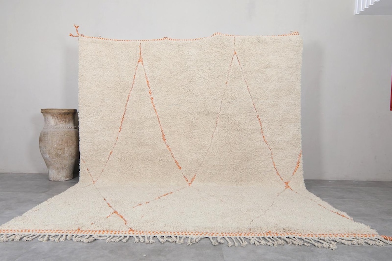 May include: Cream-colored, hand-woven rug with a geometric design. The rug features three large, orange-red diamond shapes. The edges are finished with a fringe. A decorative vase is visible in the background.