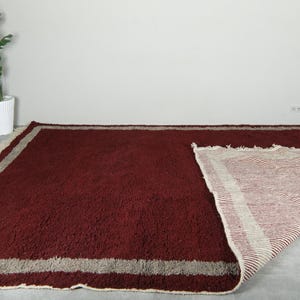 May include: A large, rectangular burgundy rug with a thick pile. The rug has a light beige border and fringe. Part of the rug is folded back, revealing a striped underside. The rug is placed on a gray floor.