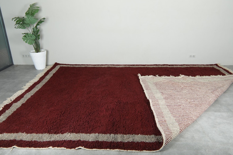 May include: A large, rectangular burgundy rug with a thick pile. The rug has a light beige border and fringe. Part of the rug is folded back, revealing a striped underside. The rug is placed on a gray floor.