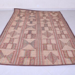 May include: A rectangular woven mat with a geometric pattern. The mat features alternating vertical stripes and geometric shapes in shades of brown, red, and beige. The border is a darker brown.