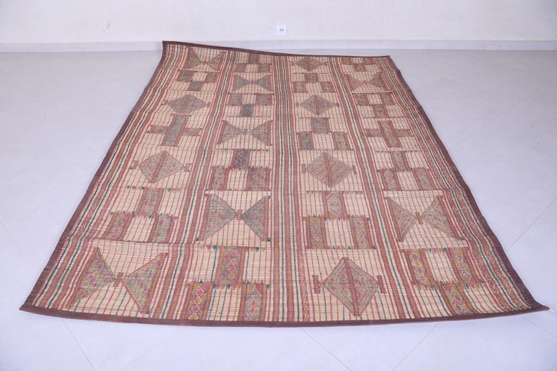 May include: A rectangular woven mat with a geometric pattern. The mat features alternating vertical stripes and geometric shapes in shades of brown, red, and beige. The border is a darker brown.
