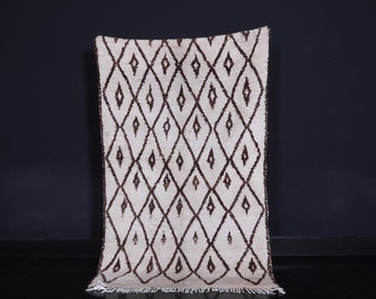 May include: A white and brown Moroccan rug with a diamond pattern. The rug has a fringe border.