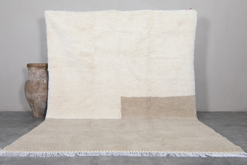 Puede incluir: Una alfombra tejida a mano de color blanco y beige con un dise&ntilde;o geom&eacute;trico. La alfombra tiene un borde de flecos.