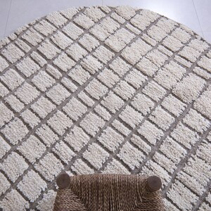 May include: A round, cream-colored rug with a geometric pattern of squares. The rug has a woven texture and is made of natural fibers.