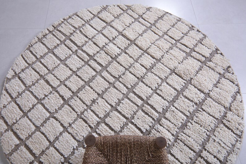 May include: A round, cream-colored rug with a geometric pattern of squares. The rug has a woven texture and is made of natural fibers.