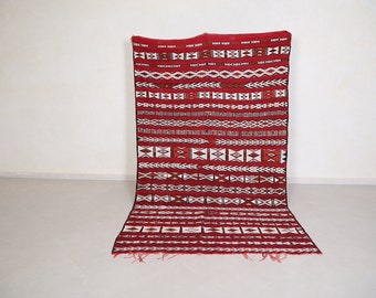 Op de afbeelding: Een rood-wit Marokkaans tapijt met ingewikkelde geometrische patronen. Het tapijt heeft een franje rand en is gemaakt van wol.