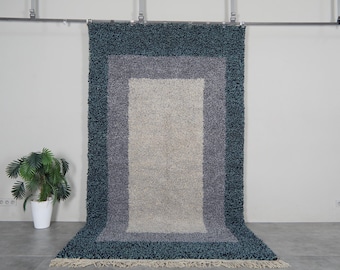 May include: A rectangular rug with a layered design in shades of teal, gray, and beige. The rug has a shaggy texture and a fringed edge. The rug is hanging against a white wall.