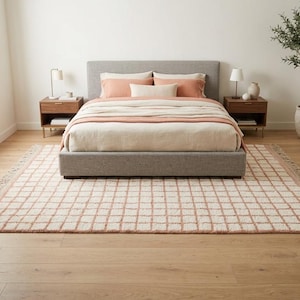 Puede incluir: Una alfombra rectangular con un patrón de cuadrícula de finas líneas color terracota sobre un fondo crema con bordes deshilachados, colocada sobre un suelo de madera clara bajo una estructura de cama gris tapizada con ropa de cama en crema y terracota.