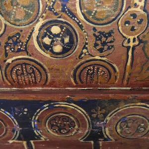 May include: Close-up of a section of a painted ceiling with intricate designs. The artwork features circular and floral motifs in shades of brown, gold, blue, and white. The style suggests a historical or cultural context, possibly from an ancient civilization.