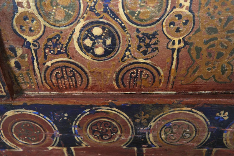 May include: Close-up of a section of a painted ceiling with intricate designs. The artwork features circular and floral motifs in shades of brown, gold, blue, and white. The style suggests a historical or cultural context, possibly from an ancient civilization.
