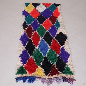 May include: A colorful Moroccan rug with a diamond pattern in shades of red, green, blue, purple, yellow, and black. The rug has a white background and a fringe border.