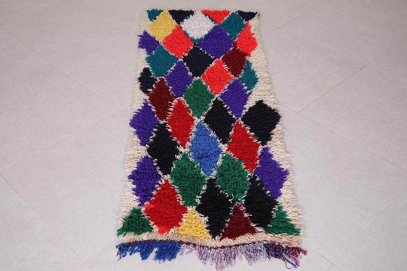 May include: A colorful Moroccan rug with a diamond pattern in shades of red, green, blue, purple, yellow, and black. The rug has a white background and a fringe border.