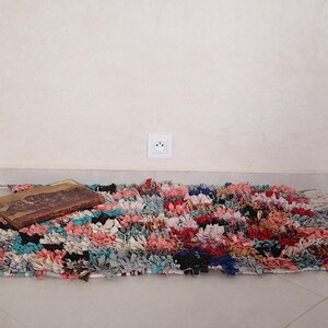 Puede incluir: Una alfombra colorida tejida a mano con una textura desgastada y deshilachada. La alfombra est&aacute; hecha de varios tonos de rosa, azul, rojo y negro. Un libro vintage est&aacute; sobre la alfombra.