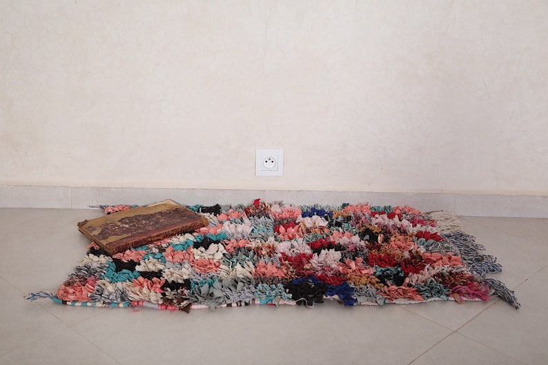Puede incluir: Una alfombra colorida tejida a mano con una textura desgastada y deshilachada. La alfombra est&aacute; hecha de varios tonos de rosa, azul, rojo y negro. Un libro vintage est&aacute; sobre la alfombra.