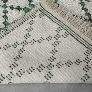 K&ouml;nnte beinhalten: Nahaufnahme eines cremefarbenen Teppichs mit einem geometrischen Muster in Gr&uuml;n und Schwarz. Der Teppich hat an einer Kante einen Fransen und eine gewebte Textur. Das Design zeigt sich wiederholende Rautenformen.