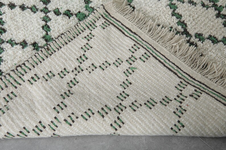 K&ouml;nnte beinhalten: Nahaufnahme eines cremefarbenen Teppichs mit einem geometrischen Muster in Gr&uuml;n und Schwarz. Der Teppich hat an einer Kante einen Fransen und eine gewebte Textur. Das Design zeigt sich wiederholende Rautenformen.