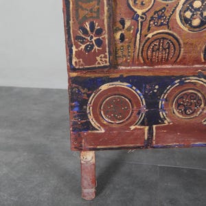 May include: Detail of a wooden chest with hand-painted patterns in red, blue, and gold. The design includes floral and geometric motifs. The chest's leg is visible. The chest is likely an antique.