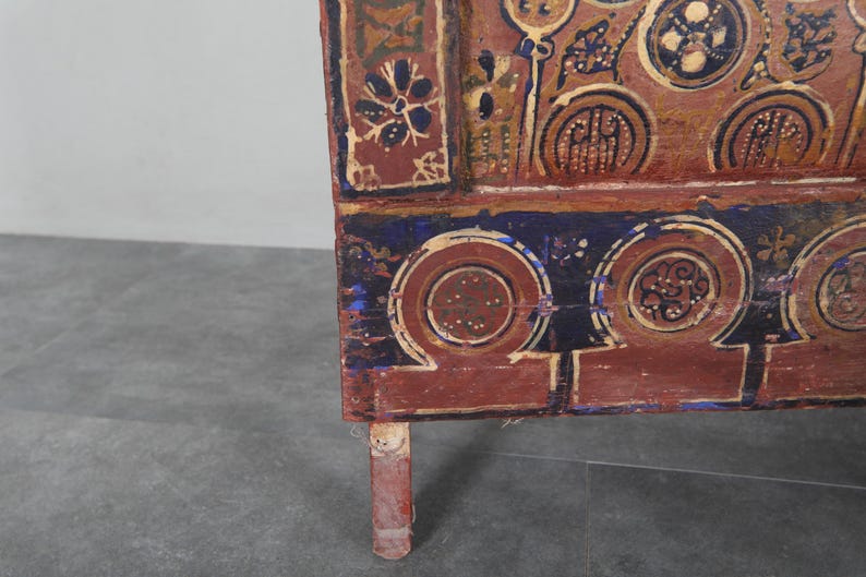May include: Detail of a wooden chest with hand-painted patterns in red, blue, and gold. The design includes floral and geometric motifs. The chest's leg is visible. The chest is likely an antique.