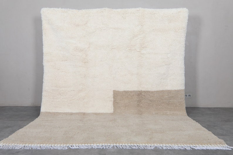 Puede incluir: Una alfombra tejida a mano de color blanco y beige con un dise&ntilde;o geom&eacute;trico. La alfombra tiene un borde de flecos.