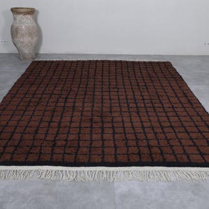 Peut inclure: Un tapis rectangulaire marron avec un motif quadrill&eacute; noir. Le tapis a une bordure noire et une frange blanche le long du bord inf&eacute;rieur. Un vase antique est en arri&egrave;re-plan.