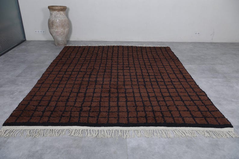 Peut inclure: Un tapis rectangulaire marron avec un motif quadrill&eacute; noir. Le tapis a une bordure noire et une frange blanche le long du bord inf&eacute;rieur. Un vase antique est en arri&egrave;re-plan.