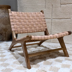 May include: A woven leather chair with a light brown leather seat and back. The chair has a wooden frame with a dark brown finish. The chair is sitting on a white rug with a geometric pattern.