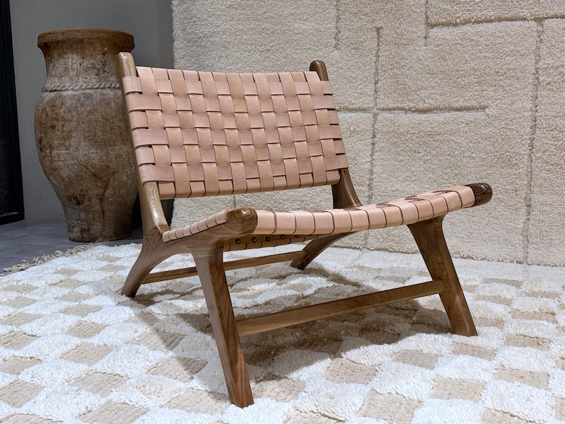May include: A woven leather chair with a light brown leather seat and back. The chair has a wooden frame with a dark brown finish. The chair is sitting on a white rug with a geometric pattern.