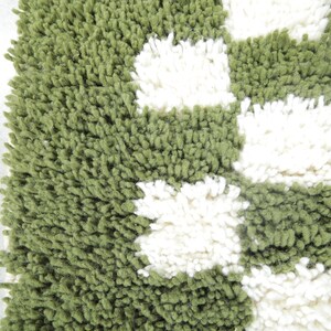 May include: Close-up of a green and white shag rug. The rug has a textured, plush appearance with a pattern of white, square-shaped accents against a green background. The rug's design is simple and modern.