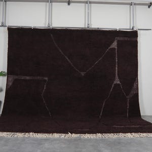 May include: A large, dark brown rug with a geometric design in lighter brown. The rug has a thick pile and a fringed edge. The design features angular shapes. The rug is displayed against a white wall.