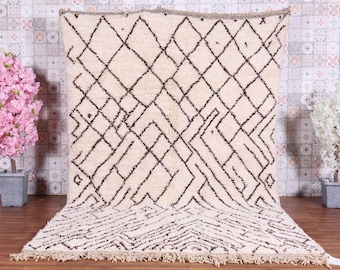 May include: Cream-colored rug with a geometric diamond pattern in black. The rug has a thick pile and a fringed edge. The design features a series of interconnected diamonds and linear shapes. The rug is displayed against a patterned wall with pink and white flowering plants on either side.