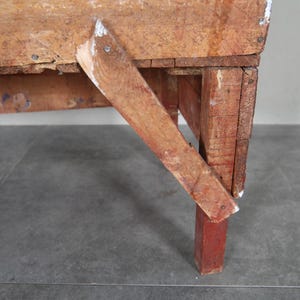 May include: Close-up of a rustic wooden table with a distressed finish. The table features a red-painted leg and a supporting brace. The wood shows signs of wear and age, with visible nail heads and paint chips.
