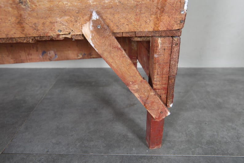May include: Close-up of a rustic wooden table with a distressed finish. The table features a red-painted leg and a supporting brace. The wood shows signs of wear and age, with visible nail heads and paint chips.