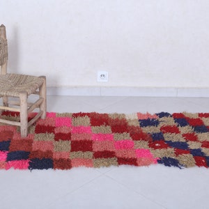 May include: A colourful, hand-woven rug with a chequered pattern in shades of pink, red, blue, and beige. The rug is made of soft, fluffy fibres and has a slightly distressed look. A small wooden chair with a woven seat sits on the rug.