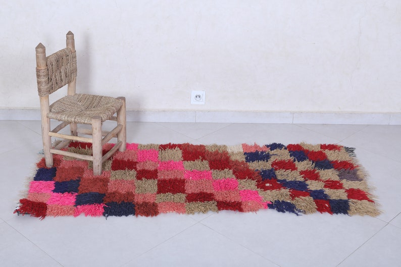 May include: A colourful, hand-woven rug with a chequered pattern in shades of pink, red, blue, and beige. The rug is made of soft, fluffy fibres and has a slightly distressed look. A small wooden chair with a woven seat sits on the rug.