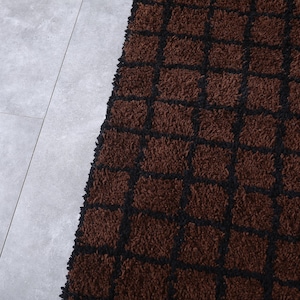 Peut inclure: Gros plan sur un tapis marron avec un motif quadrill&eacute; noir. Le tapis a une texture touffue et est pos&eacute; sur un sol carrel&eacute; gris clair. La couleur du tapis est un brun riche et chaud.
