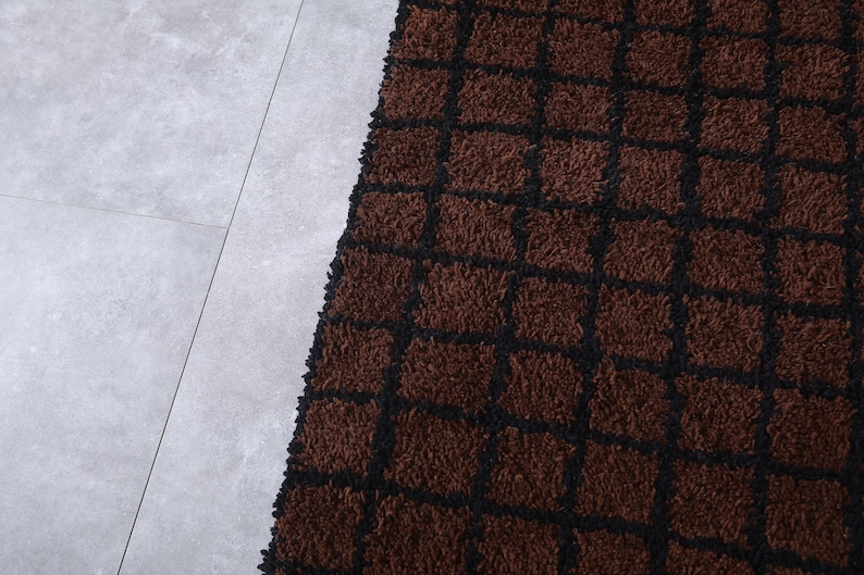 Peut inclure: Gros plan sur un tapis marron avec un motif quadrill&eacute; noir. Le tapis a une texture touffue et est pos&eacute; sur un sol carrel&eacute; gris clair. La couleur du tapis est un brun riche et chaud.