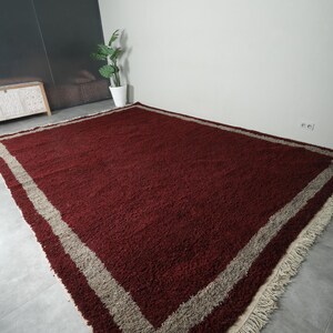 May include: A large, rectangular rug with a deep burgundy central field. A wide, light beige border frames the rug, and the edges feature a fringe. The rug is made of a thick, textured material.