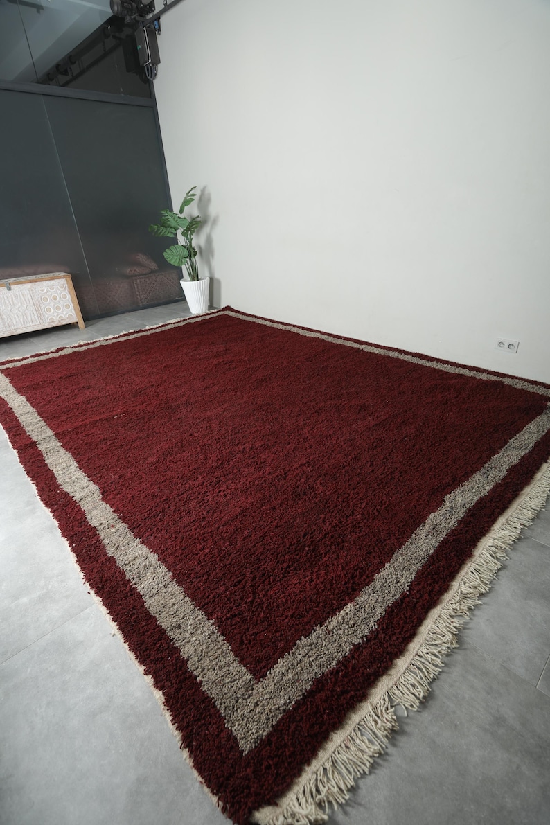 May include: A large, rectangular rug with a deep burgundy central field. A wide, light beige border frames the rug, and the edges feature a fringe. The rug is made of a thick, textured material.
