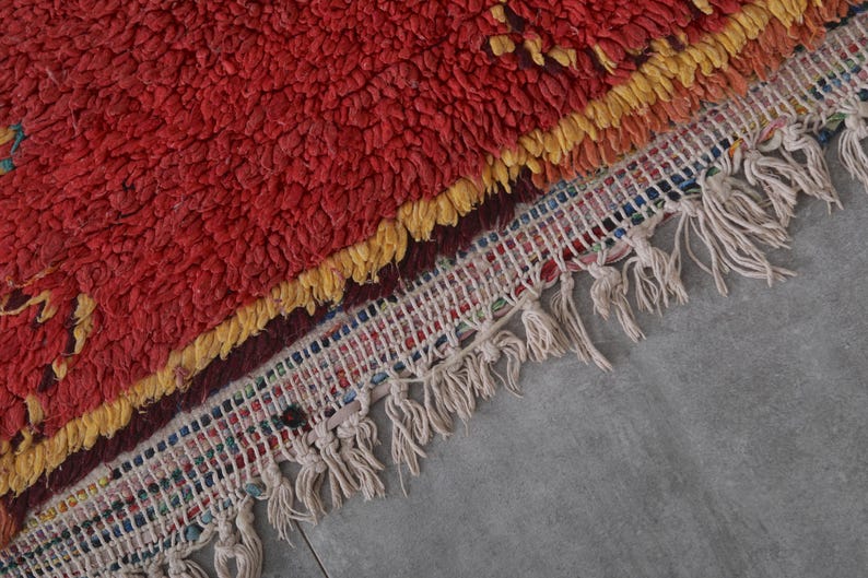 May include: A red, yellow, and white Moroccan rug with a fringe border. The rug has a textured, fluffy surface.