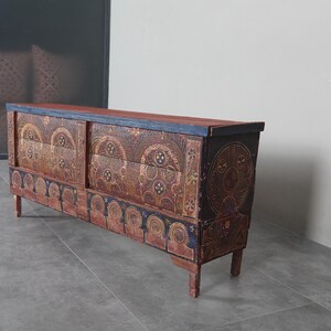 May include: A long wooden chest with detailed carved patterns in brown, red, and blue. It has a flat top and four short legs. The front has sliding doors with circular and arched designs. The chest is likely made of wood.
