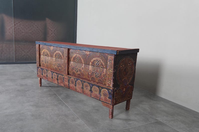 May include: A long wooden chest with detailed carved patterns in brown, red, and blue. It has a flat top and four short legs. The front has sliding doors with circular and arched designs. The chest is likely made of wood.