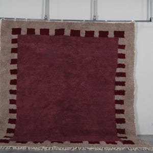 May include: A large, rectangular rug with a deep burgundy center and a tan border. The border features a repeating pattern of burgundy rectangles. The rug has a shaggy texture and a fringe along the bottom edge. It is displayed against a white wall.
