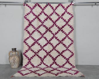 May include: A white and purple Moroccan rug with a diamond pattern. The rug is handwoven and has a soft, fluffy texture.