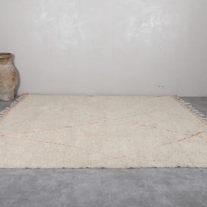 May include: Cream-colored, rectangular rug with a geometric design. The rug features a soft, textured surface and orange accent lines. The edges are finished with a fringe detail. A decorative, beige vase is in the background.
