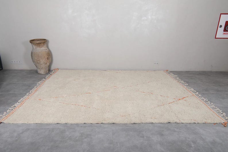 May include: Cream-colored, rectangular rug with a geometric design. The rug features a soft, textured surface and orange accent lines. The edges are finished with a fringe detail. A decorative, beige vase is in the background.