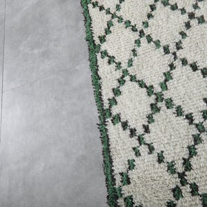 K&ouml;nnte beinhalten: Nahaufnahme eines cremefarbenen Teppichs mit einem geometrischen Rautenmuster in Dunkelgr&uuml;n. Der Teppich hat eine strukturierte Oberfl&auml;che und einen gr&uuml;nen Fransenrand. Der Teppich liegt auf einer grauen Oberfl&auml;che.