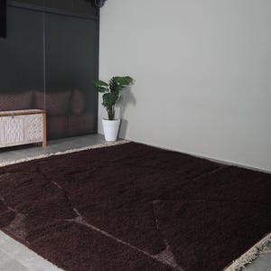 May include: A large, rectangular, dark brown rug with a shaggy texture. The rug has a cream-colored fringe along the edges and lighter brown geometric patterns. It is placed on a gray concrete floor.