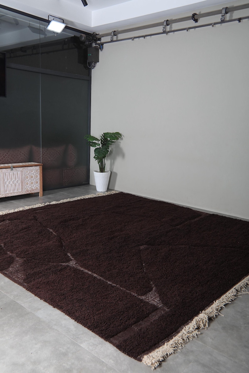 May include: A large, rectangular, dark brown rug with a shaggy texture. The rug has a cream-colored fringe along the edges and lighter brown geometric patterns. It is placed on a gray concrete floor.